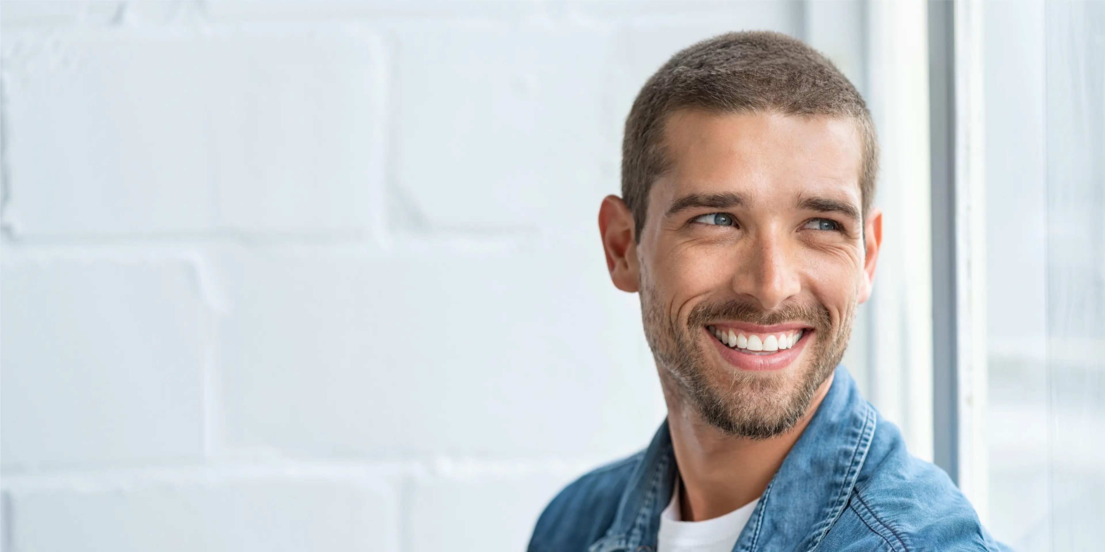 Smiling man with short hair and beard looking out window wearing a blue denim jacket. La Perle Dental Center in Edmonton, AB TST 3T9