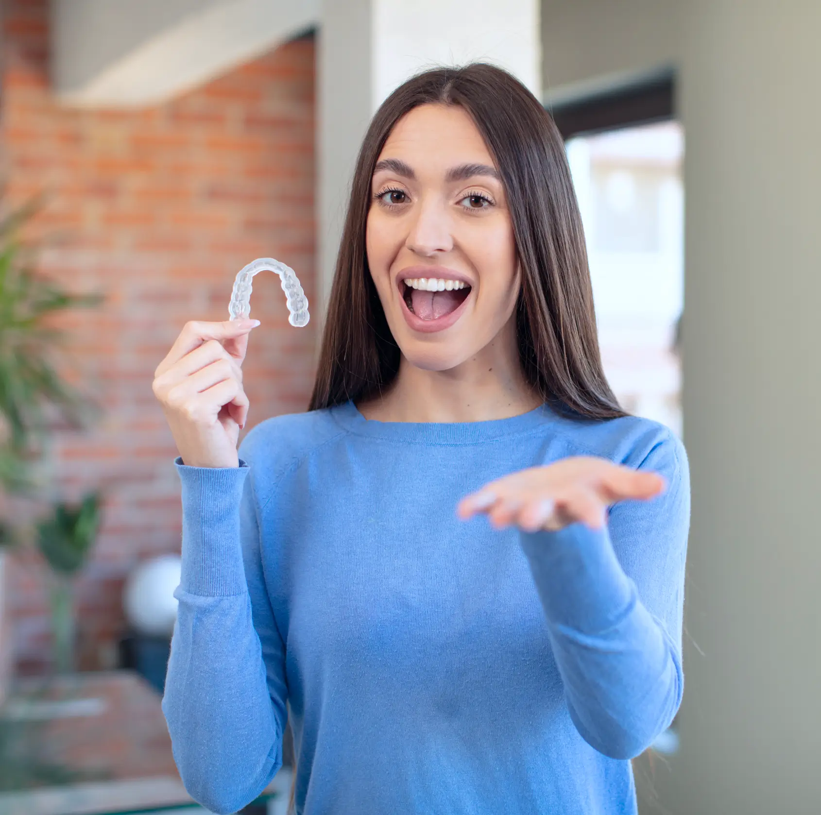 Smiling woman in blue sweater holding a clear dental aligner for teeth straightening. La Perle Dental Center in Edmonton, AB TST 3T9