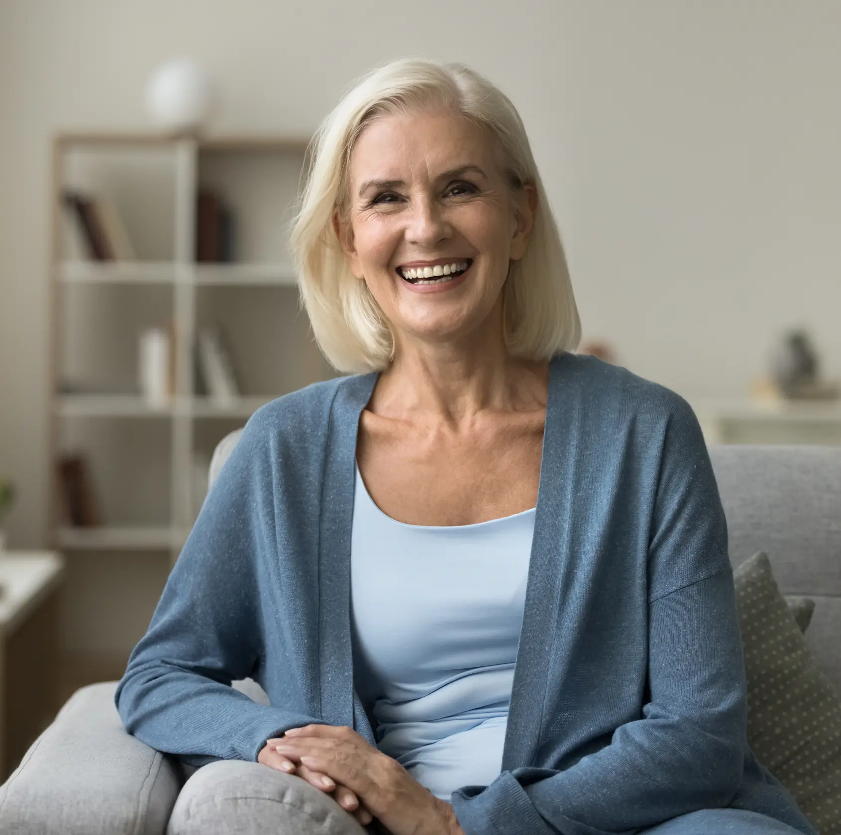 Smiling elderly woman with blonde hair sitting on a gray couch in a light room. La Perle Dental Center in Edmonton, AB TST 3T9
