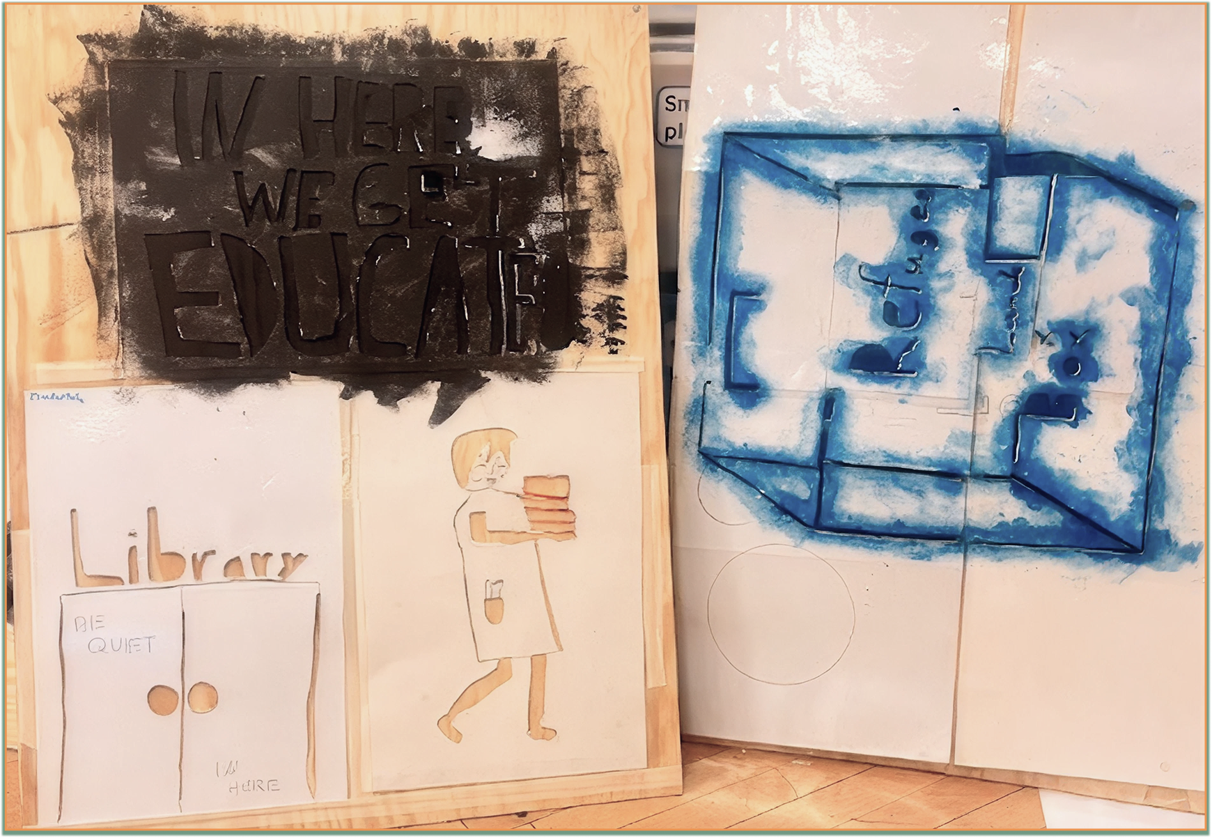 Wooden panel art with black stencil text 'IN HERE WE GET EDUCATED' above two white panels; the left panel has the word 'Library' above doors labeled 'BE QUIET' and 'I AM HERE'; the right panel shows a simple drawing of a person carrying stacked books. Next to these panels is a white board with a blue spray-painted blueprint labeled 'Library' with an 'X' marking a spot.