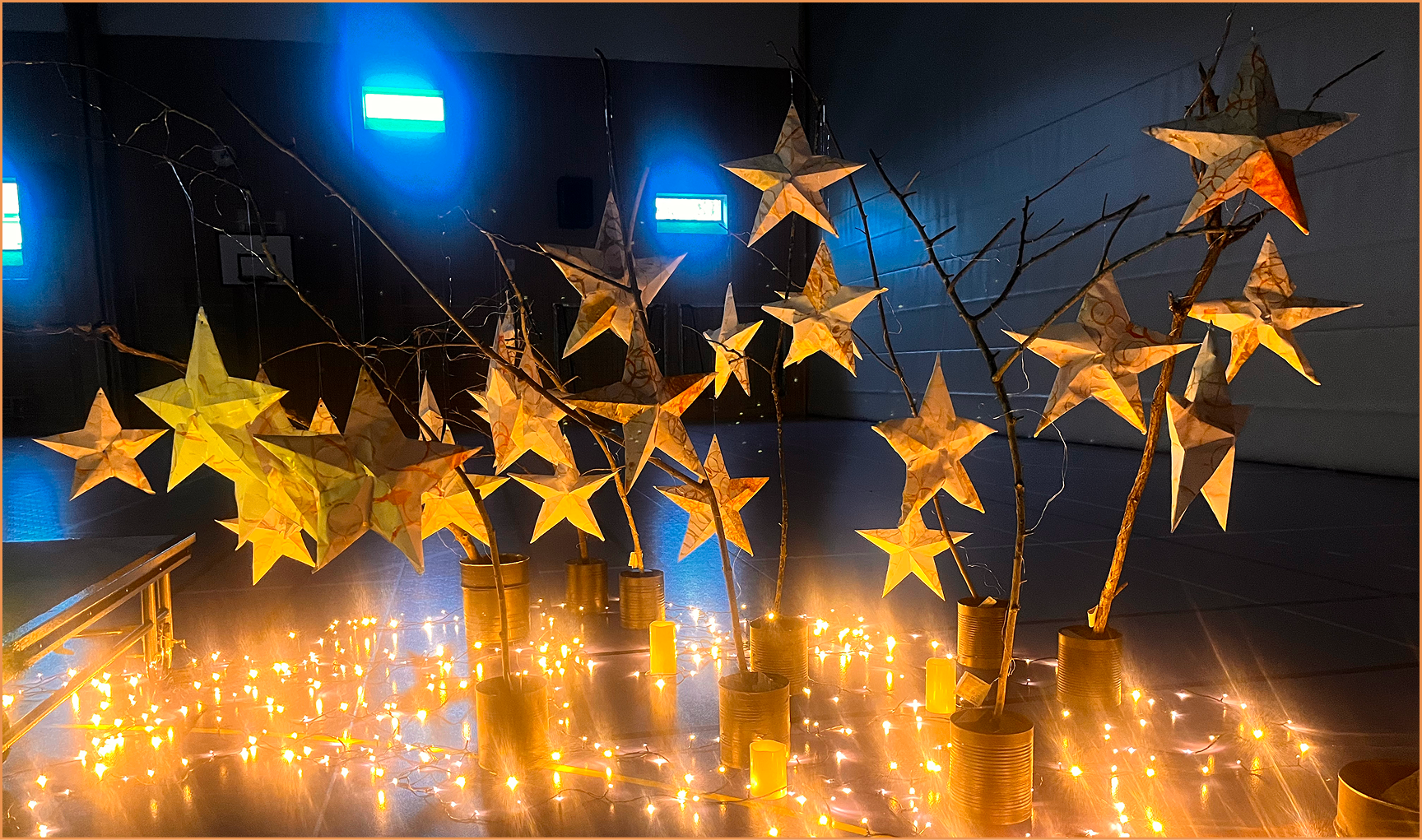 Decorative display of paper star lanterns hanging from bare tree branches placed in tin cans, surrounded by glowing string lights and small candles on the floor in a dimly lit indoor space.