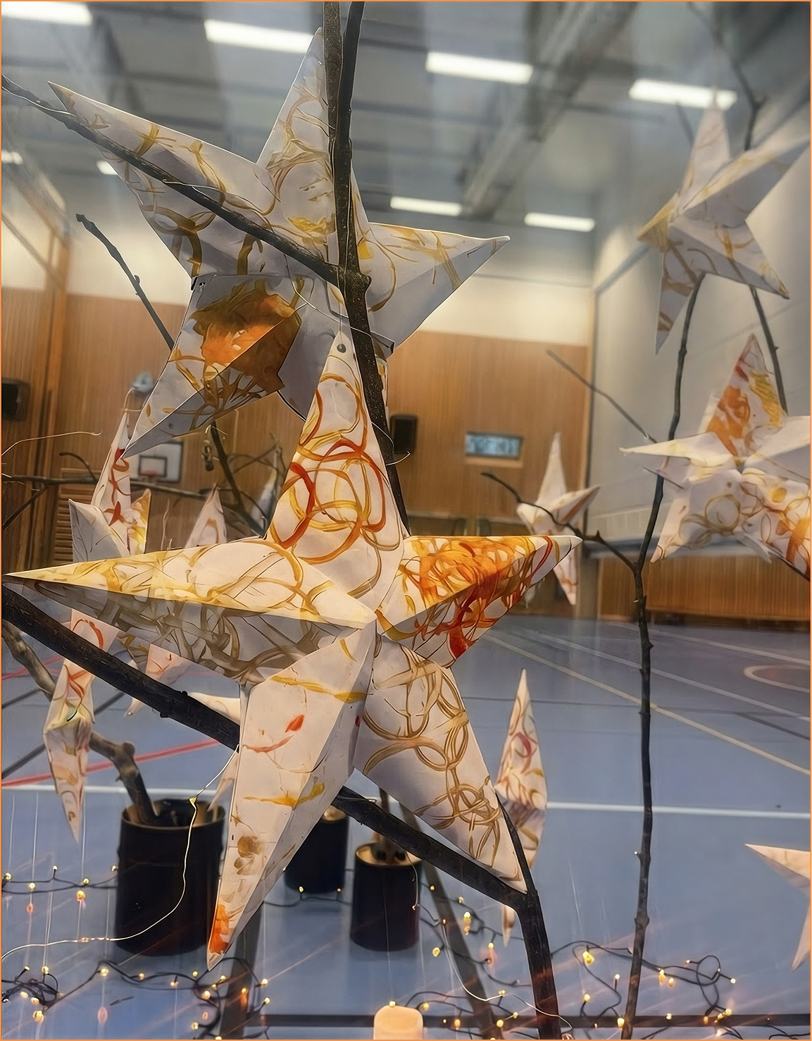 Decorative paper stars with orange and yellow swirls hanging on black branches in a gymnasium with a basketball hoop in the background.