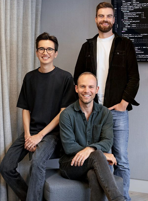 Trois hommes souriants posant dans un bureau moderne avec un écran d'ordinateur en arrière-plan.