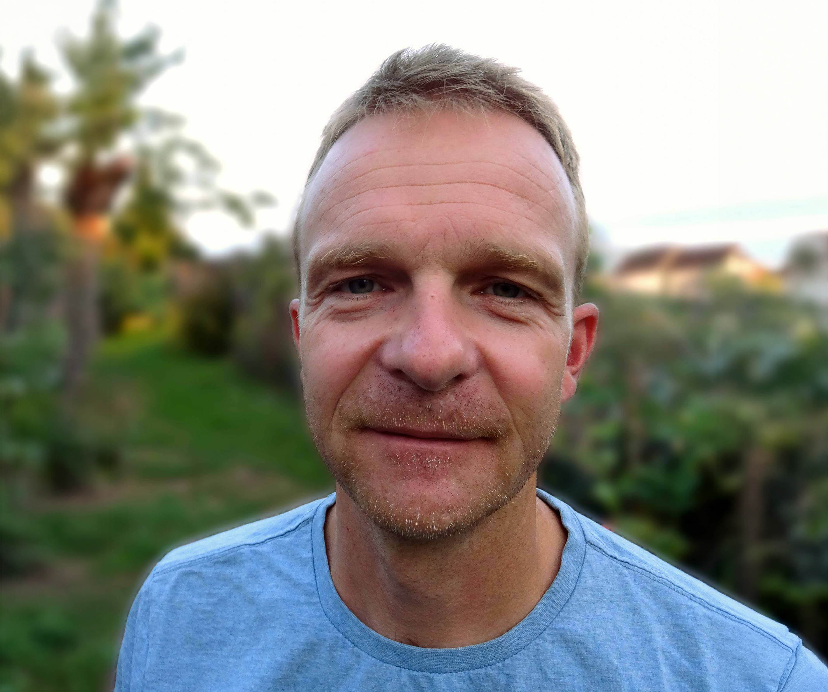 Thomas Emmenegger mit kurzen blonden Haaren und hellblauem T-Shirt in einem Garten mit Büschen und Bäumen leicht lächelnd in die Kamera blickend.