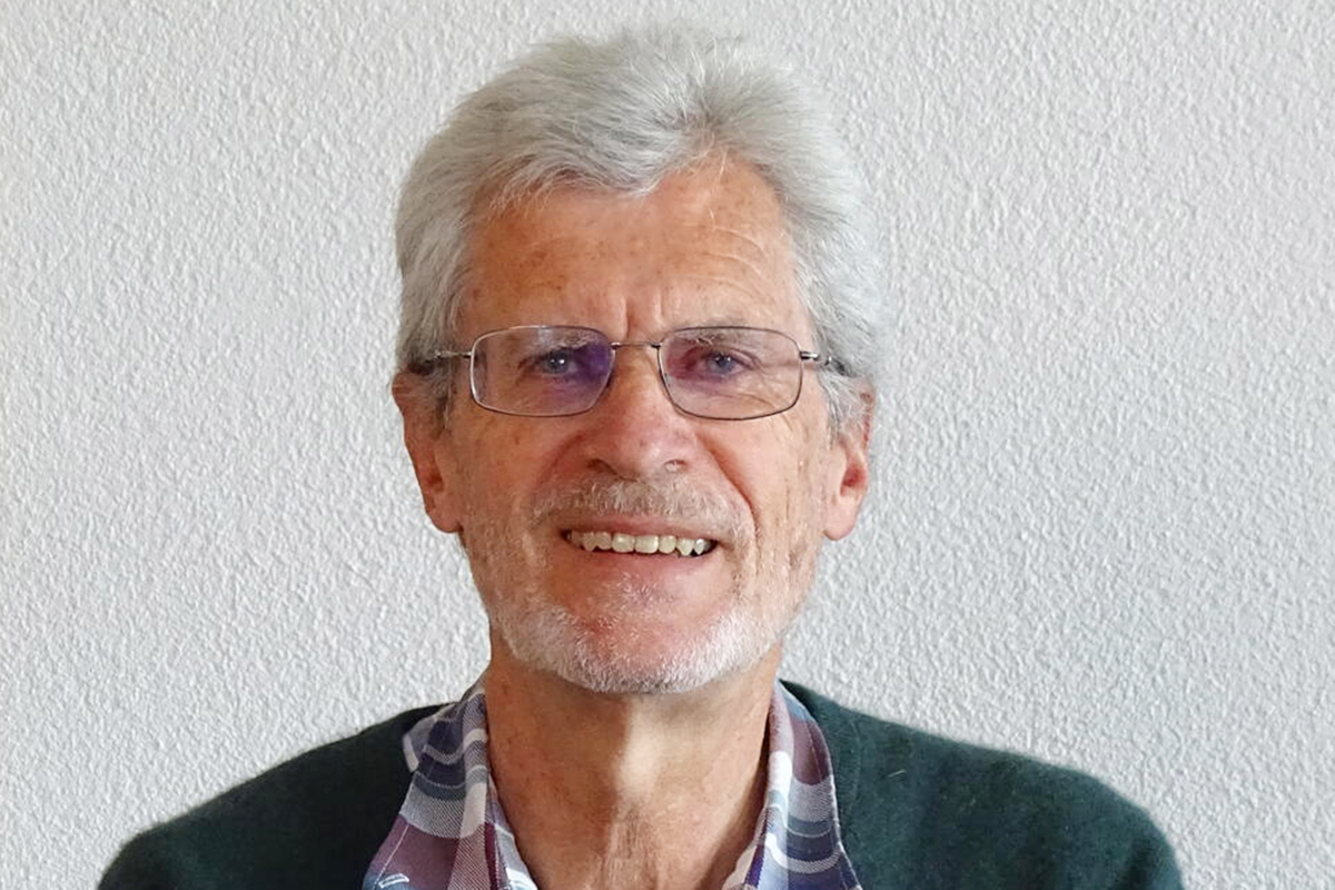 Urs Brandenberg mit grauen Haaren und schmaler Brille in dunkelgrüner Strickjacke über einem karierten Hemd vor einer weissen Wand lächelnd in die Kamera blickend.