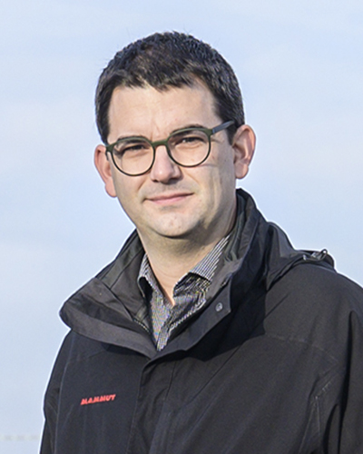 Michael Hoekstra mit runder Brille und dunkler Jacke vor hellem Himmel leicht zur Seite gedreht und in die Kamera blickend.