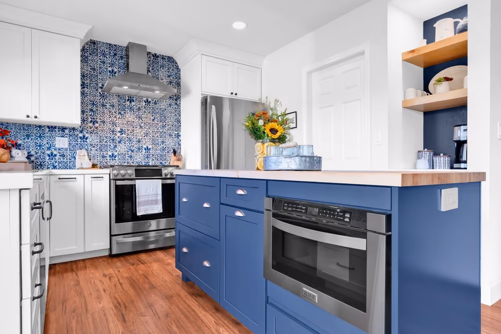 Modern kitchen with blue lower cabinets and white upper cabinets, featuring a wooden countertop island, stainless steel oven and stove with blue and white patterned tile backsplash, and wooden floor.