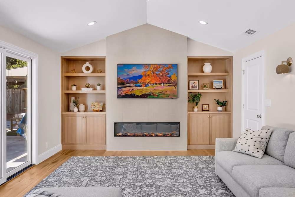 Modern living room with vaulted ceiling, light wood built-in shelves flanking a wall-mounted electric fireplace, colorful landscape painting above, gray upholstered sofa with patterned pillows on the right, and sliding glass door on the left.