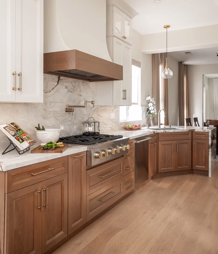 Modern kitchen with wooden lower cabinets and white upper cabinets, marble countertops and backsplash, stainless steel gas stove and dishwasher, a book stand with an open cookbook, fresh asparagus and vegetables on a cutting board, a hanging glass pendant light, and a corner sink with faucet under a window with curtains.