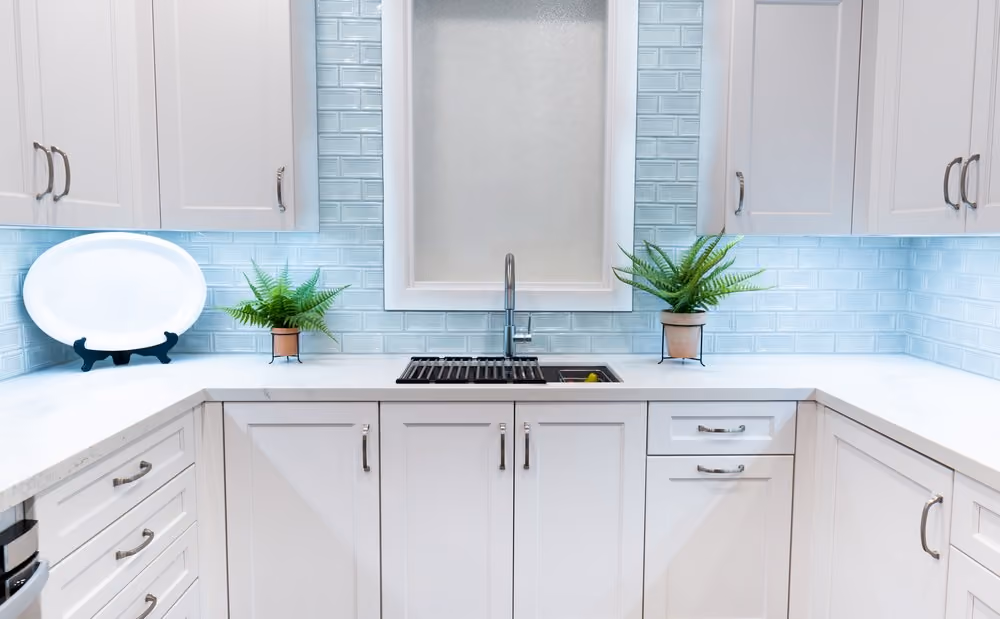Modern white kitchen with shaker style cabinets, light blue subway tile backsplash, marble countertops, stainless steel sink with faucet under a frosted window, two small potted ferns, and a decorative white plate on a black stand.