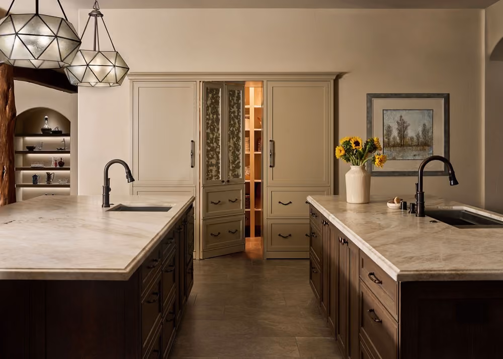 Modern kitchen with two marble-topped islands featuring dark wood cabinets, black faucets, and integrated sinks, illuminated by geometric pendant lights, with a vase of sunflowers on the right island and a partially open pantry with double doors in the background.