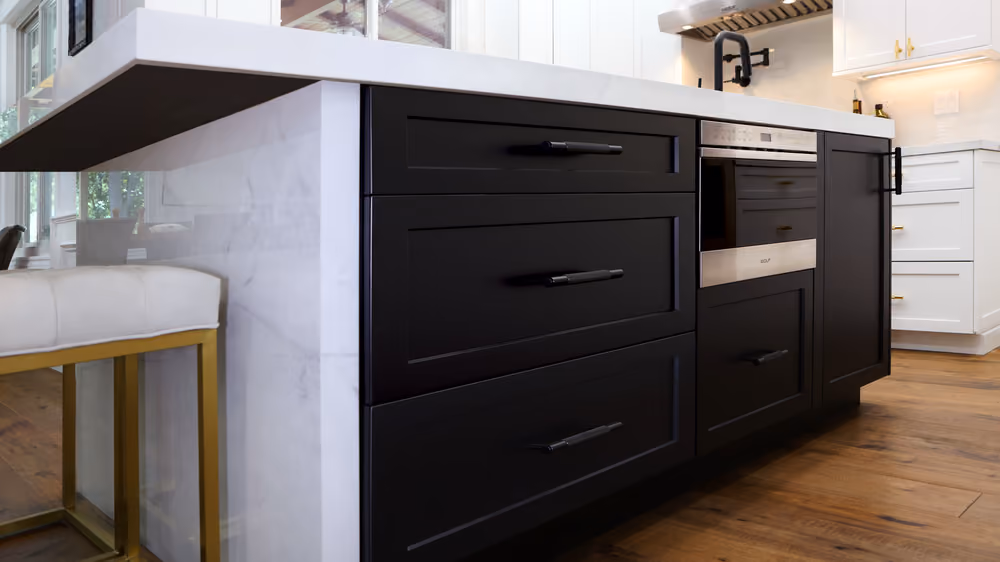 Close-up of a modern kitchen island with black cabinets, sleek metal handles, a built-in oven, and a white marble countertop, with wooden flooring and a white cushioned stool with gold legs.