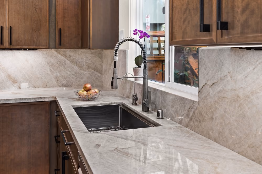Modern kitchen countertop with light gray marble surface and backsplash, stainless steel sink with high-arc spring faucet, dark wooden cabinets with black handles, a bowl of apples, and a small purple orchid plant on the windowsill.