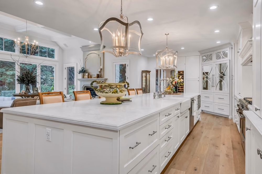 Bright modern kitchen with a large white marble island featuring multiple drawers, a farmhouse sink, and two ornate pendant lights hanging above; wooden floor and white cabinetry with mirrored glass panels; dining area with chairs and a large window showing outdoor greenery.