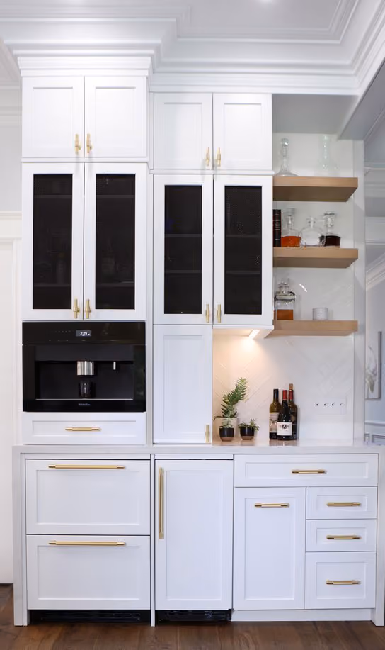 Modern kitchen cabinetry with white cabinets featuring black mesh panel doors and gold handles, a built-in black coffee machine, open wooden shelves with glass bottles, and small potted plants on the white countertop.