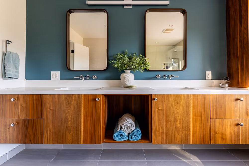 Modern bathroom vanity with double sinks and rectangular mirrors mounted on a teal accent wall, featuring a white countertop, wood cabinetry, and neatly rolled towels displayed in an open shelf below, with a vase of green plants between the sinks.