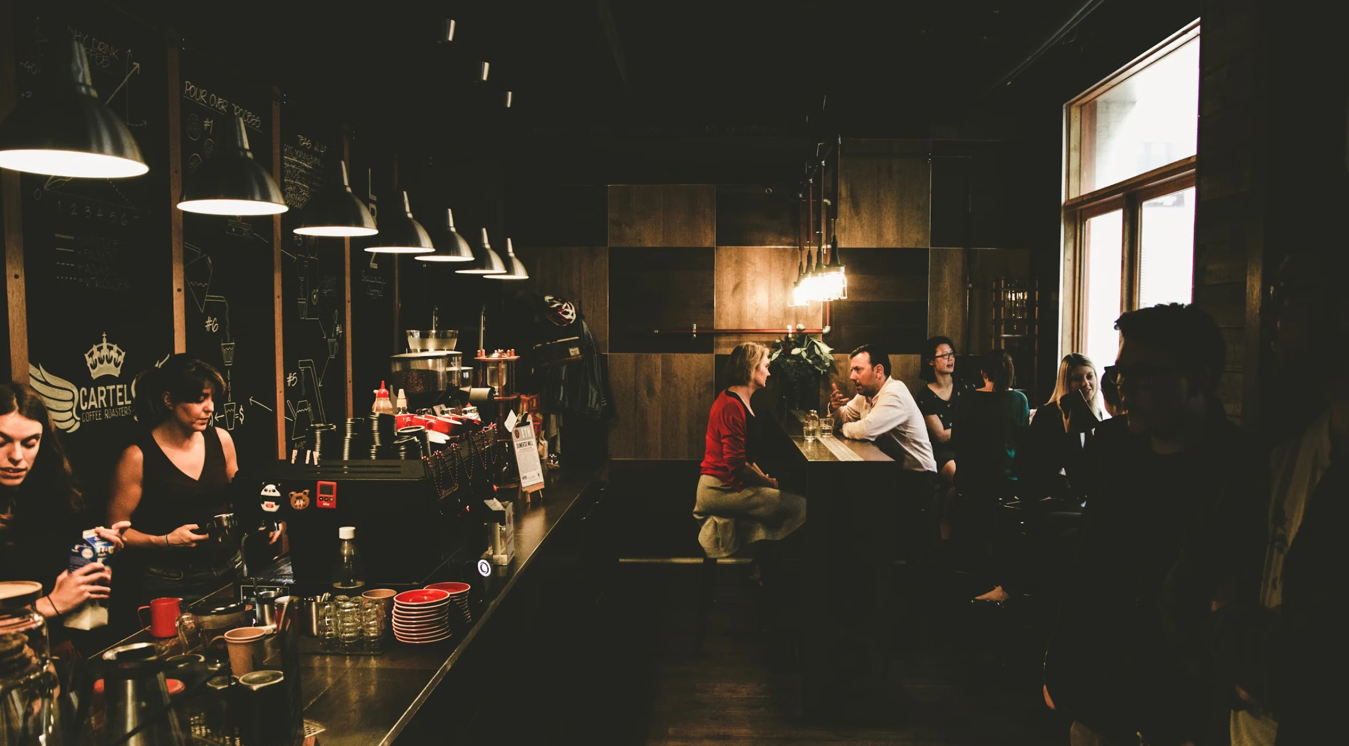 Dim coffee shop interior with baristas working and customers seated at counter and tables.
