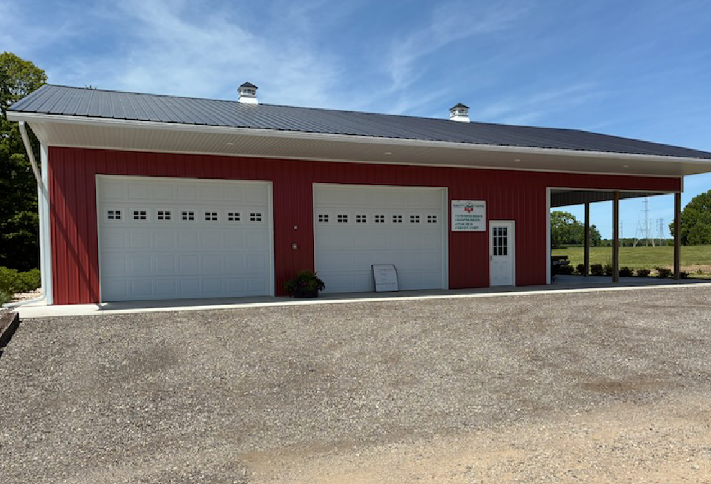 Red barn with two white garage doors, a white entry door, and a covered area on a gravel driveway.