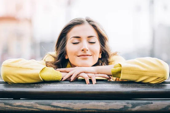 Young woman with closed eyes and a relaxed smile rests her chin on folded hands atop a wooden surface outdoors.