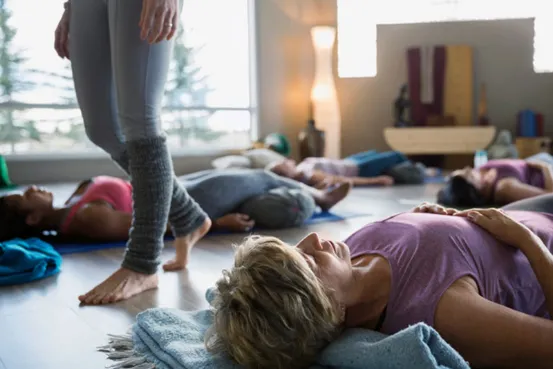 Group of people lying on yoga mats in a calm, softly lit room, with one person standing barefoot wearing leg warmers.