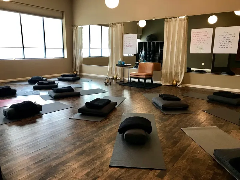 Interior of a yoga or meditation studio with dark wooden floors, several yoga mats laid out with folded blankets and pillows on each, large mirrors on one wall, beige curtains, a brown chair near a small round table, and large windows letting in natural light.