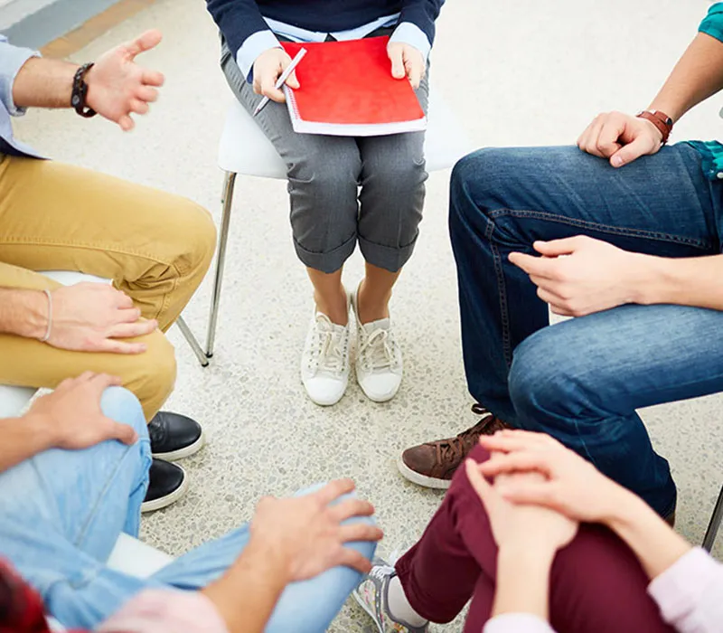 A group therapy session showing five people sitting in a circle, focusing on their legs and hands, with one person holding a red notebook and pen.