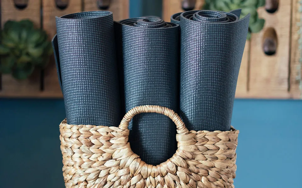 Three rolled black yoga mats standing upright inside a woven wicker basket with a circular handle, set against a blue wall with blurred wooden pegs and green plant in the background.