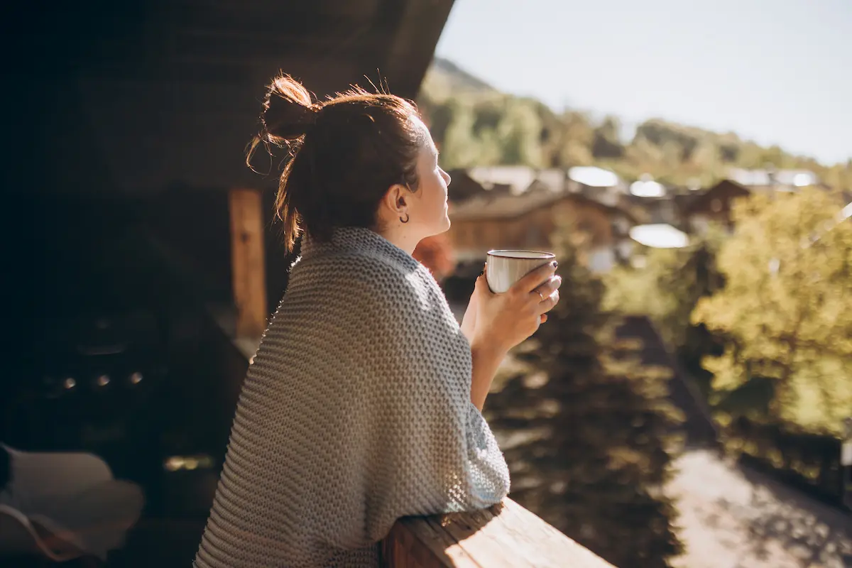 Woman wrapped in a knitted blanket standing on a wooden balcony holding a cup and looking out at a sunny, tree-filled landscape with scattered houses.
