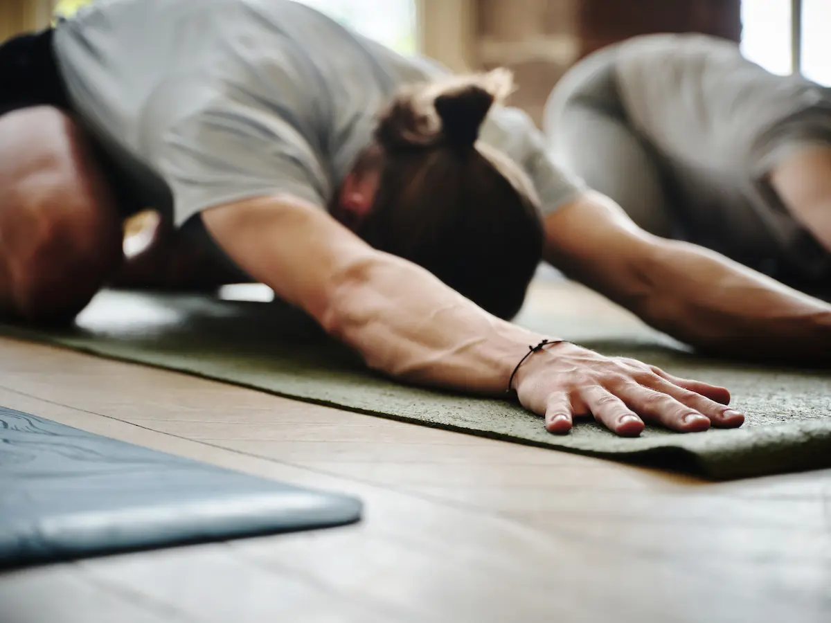 Close-up of a person with long hair tied back stretching forward on a green yoga mat indoors with another person also stretching in the background.