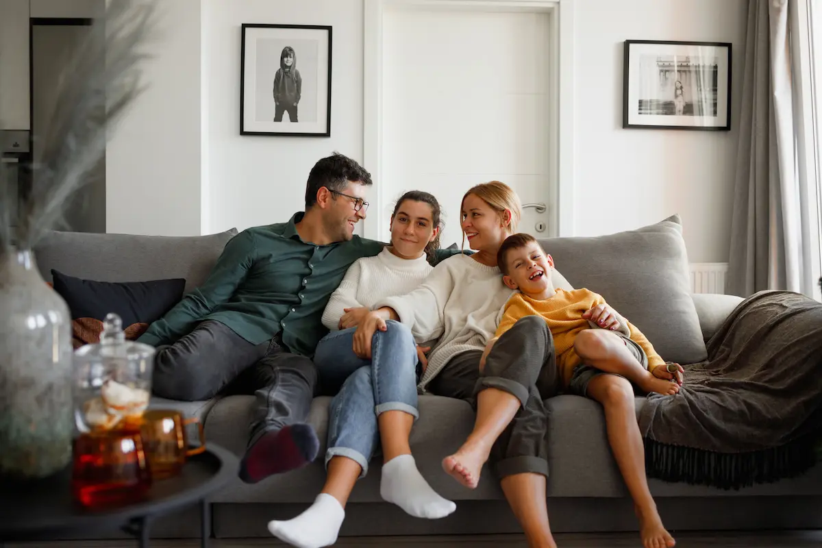 A family of four sitting close together on a gray sofa in a living room, smiling and enjoying each other's company, with two framed pictures on the wall behind them.