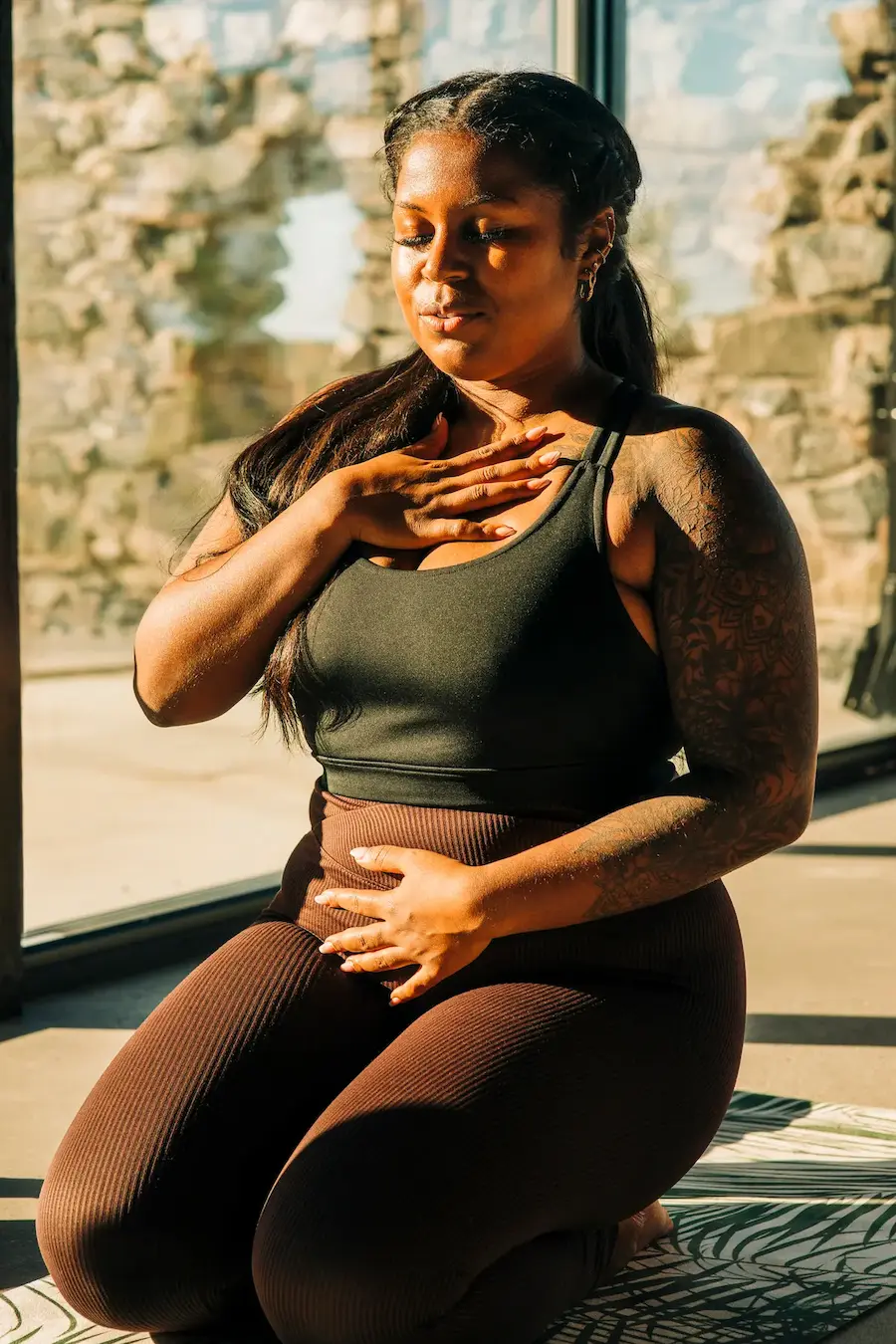 Woman with braided hair wearing a black tank top and brown leggings kneels on a patterned mat, practicing deep breathing with one hand on her chest and the other on her abdomen in a sunlit room.