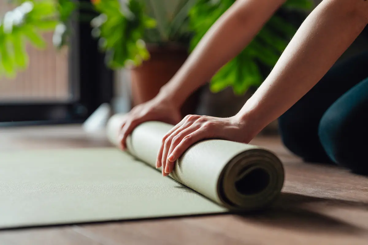 Close-up of hands rolling out a light green yoga mat on a wooden floor near a window with green plants in the background.