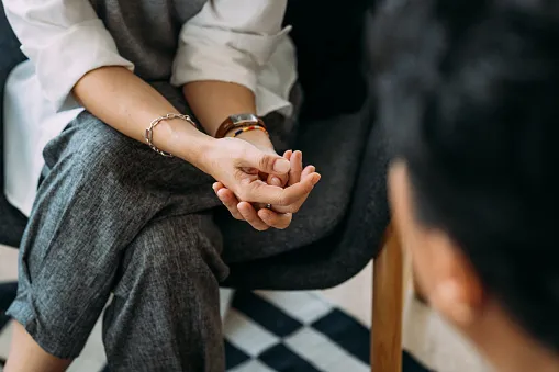 Close-up of a person sitting with legs crossed and hands gently clasped, wearing a silver bracelet and a wristwatch, in a conversation with another person partially visible in the foreground.