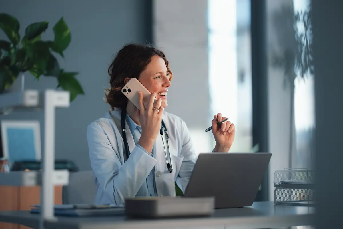 Female doctor wearing a white coat and stethoscope, smiling and talking on a smartphone while sitting at a desk with a laptop in a bright, modern office.