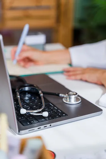 A stethoscope resting on a laptop keyboard with a person in a white coat writing in a notebook in the background.