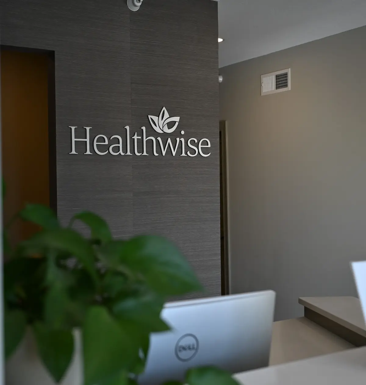 Interior office wall with the word 'Healthwise' and a leaf logo mounted on a textured dark panel behind a desk with a Dell laptop and a green leafy plant in soft focus.