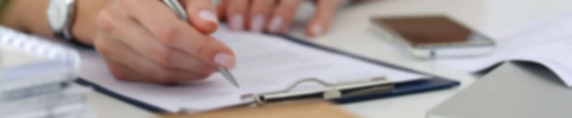 Close-up of a person's hand holding a pen and filling out a form or document on a clipboard, with a smartphone and other papers on a white desk.