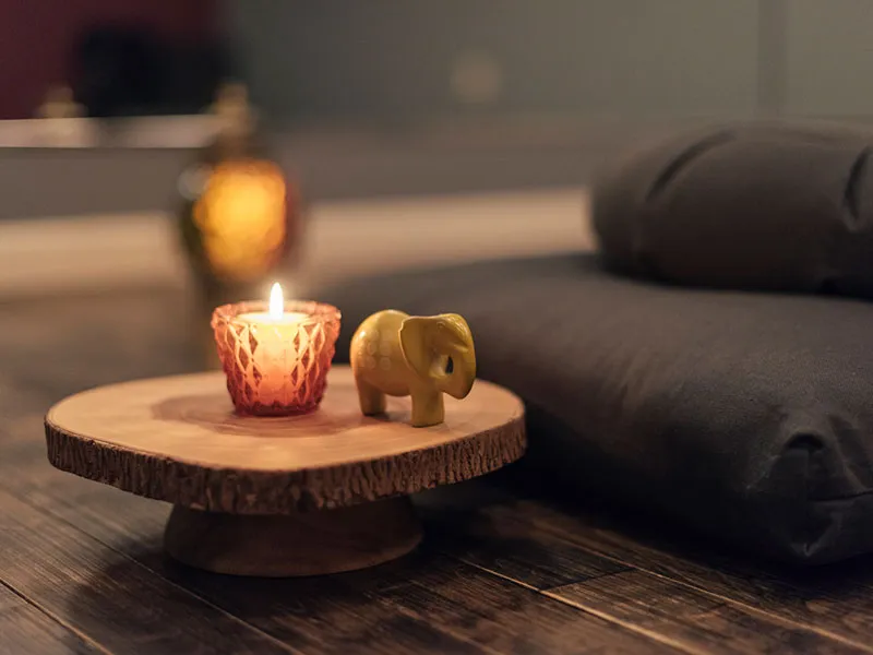 Small wooden pedestal on a wooden floor holding a lit candle in a red glass holder and a small yellow elephant figurine, next to a dark gray rolled yoga mat or cushion in a softly lit room.