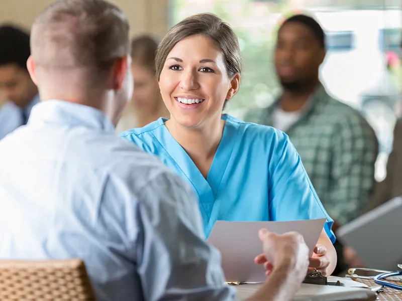 A smiling healthcare professional in blue scrubs is talking with a man sitting across the table, holding a paper.