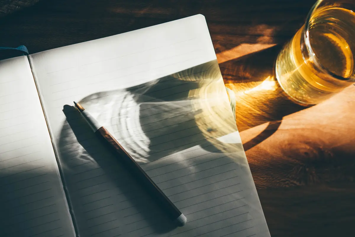 Open lined notebook on a wooden surface with a pen resting on its pages and sunlight casting shadows from a nearby glass of liquid.