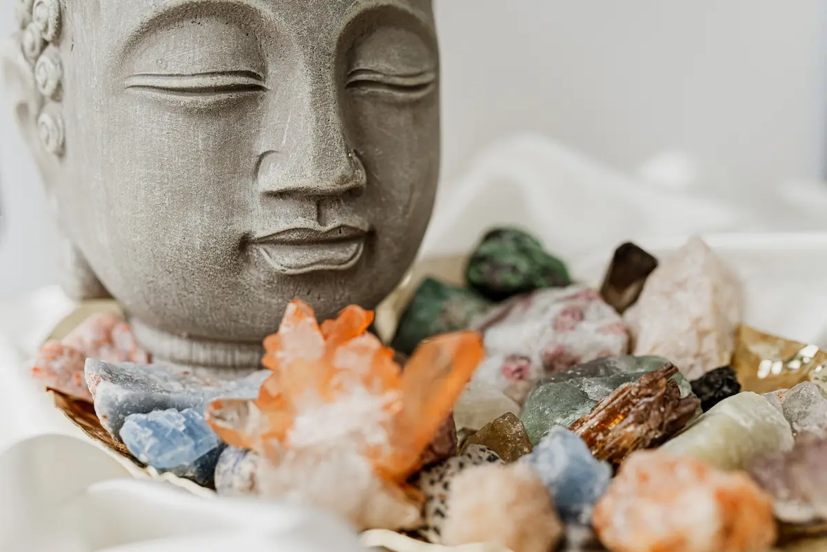 Stone Buddha head statue surrounded by various colorful crystals and stones on a soft white fabric background.