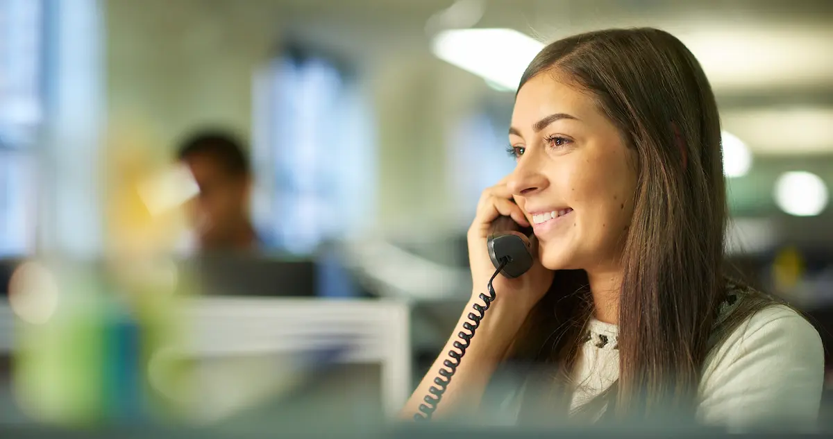 Smiling woman with long brown hair speaking on a corded telephone in a bright, modern office setting.