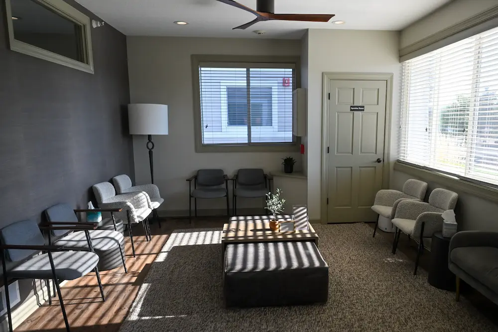 Sunlit waiting room with various chairs arranged along the walls and a central table with a small plant and books, featuring a door labeled 'Speaker Room' and large windows with blinds.