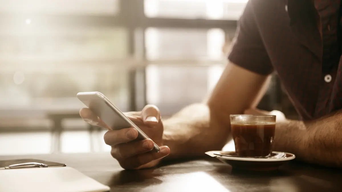 Person in a dark shirt sitting at a table holding and using a smartphone with one hand, with a glass cup of coffee on a saucer nearby and a closed notebook on the table in soft natural light.