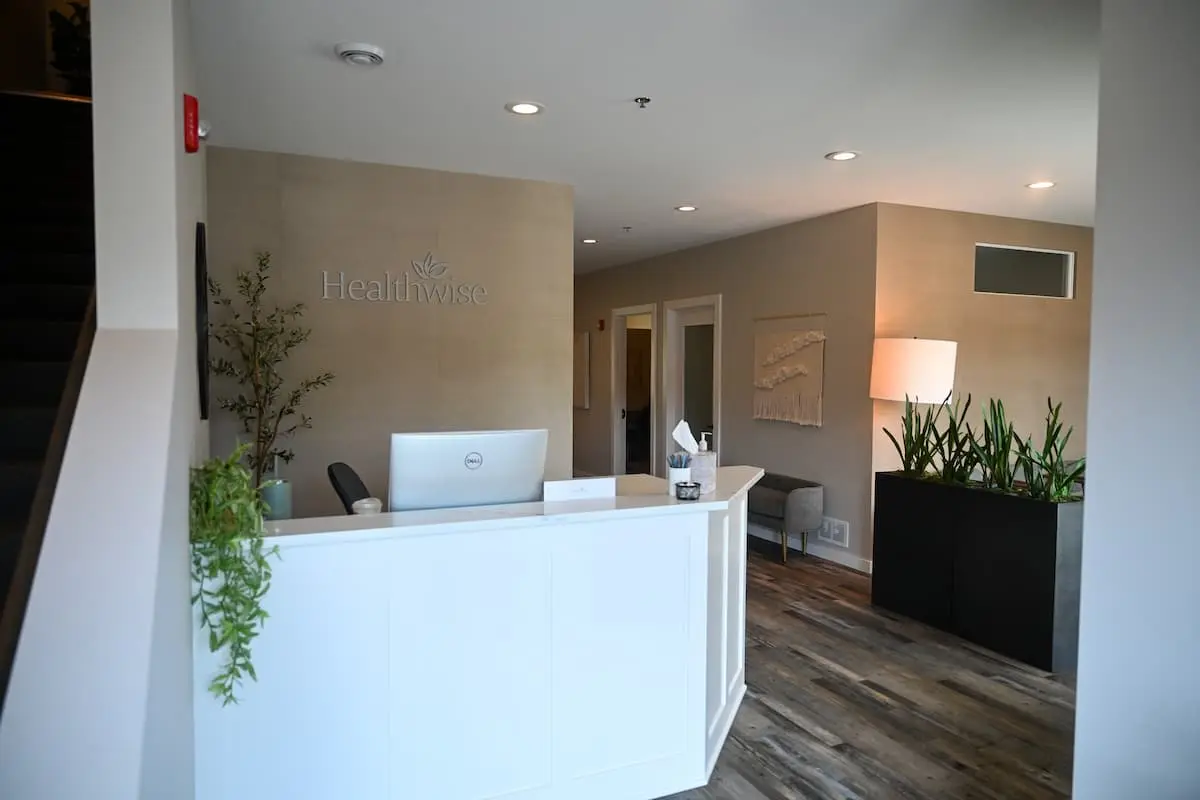 Reception area with a white desk, a Dell computer, and a plant on the left side, with a beige wall behind displaying the word Healthwise, next to a hallway with doors and a lamp on the right by tall planter boxes containing green plants.