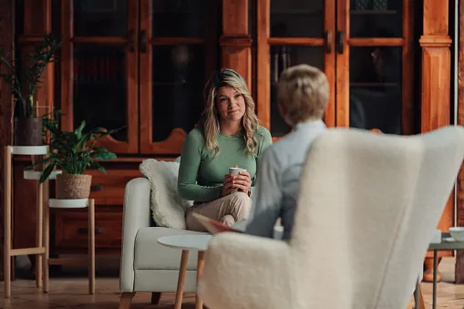 Two women engaged in a conversation in a cozy room with wooden shelves and plants, one holding a cup while sitting on a white chair.