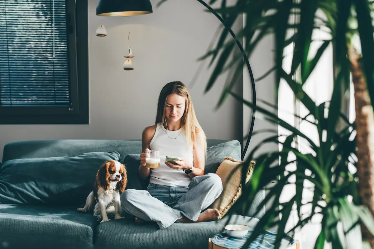 A woman sitting cross-legged on a blue couch holding a cup and looking at her smartphone, with a small dog sitting beside her.