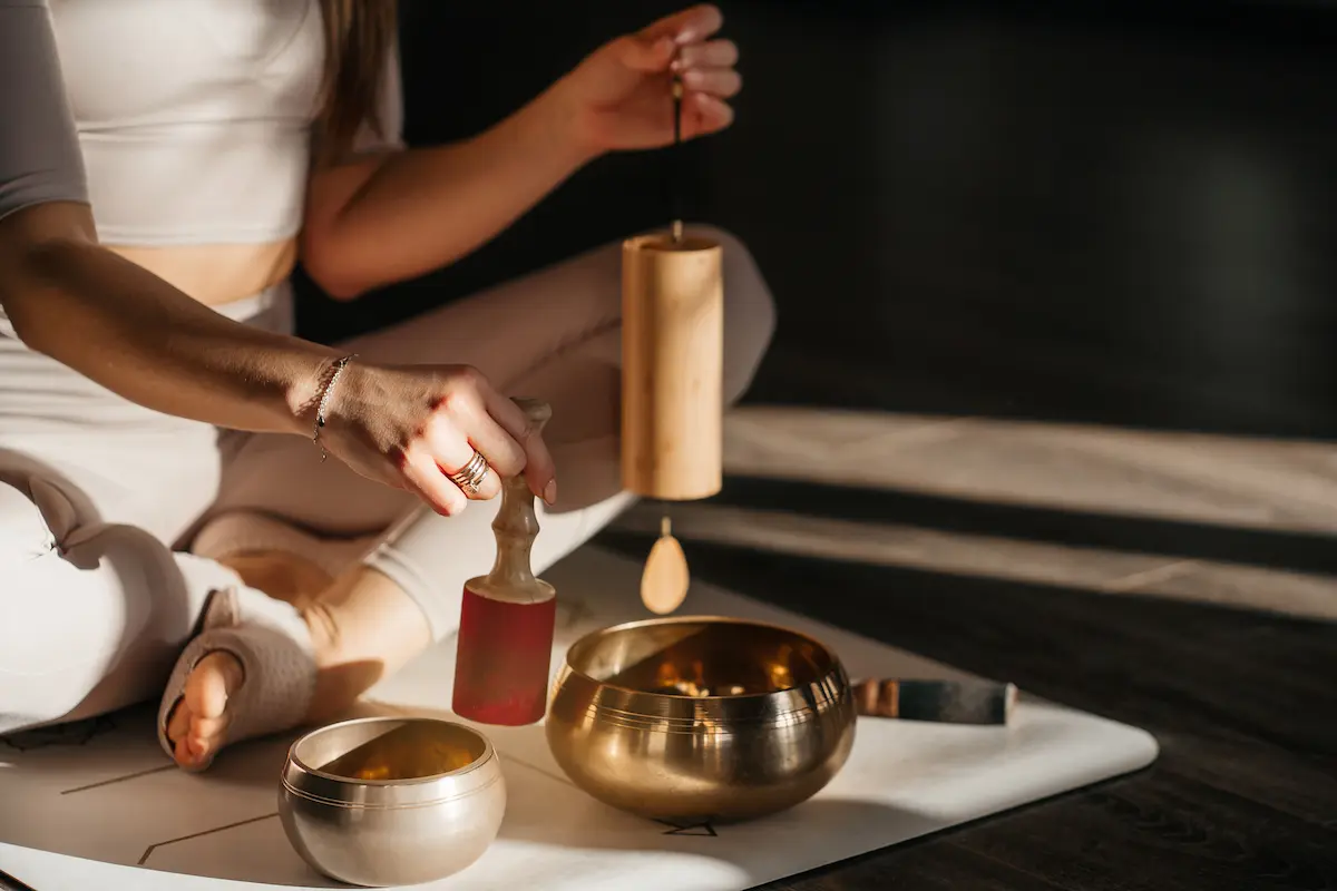 Person sitting cross-legged on a mat holding a wooden mallet and a wooden striker, playing two brass singing bowls in soft natural light.