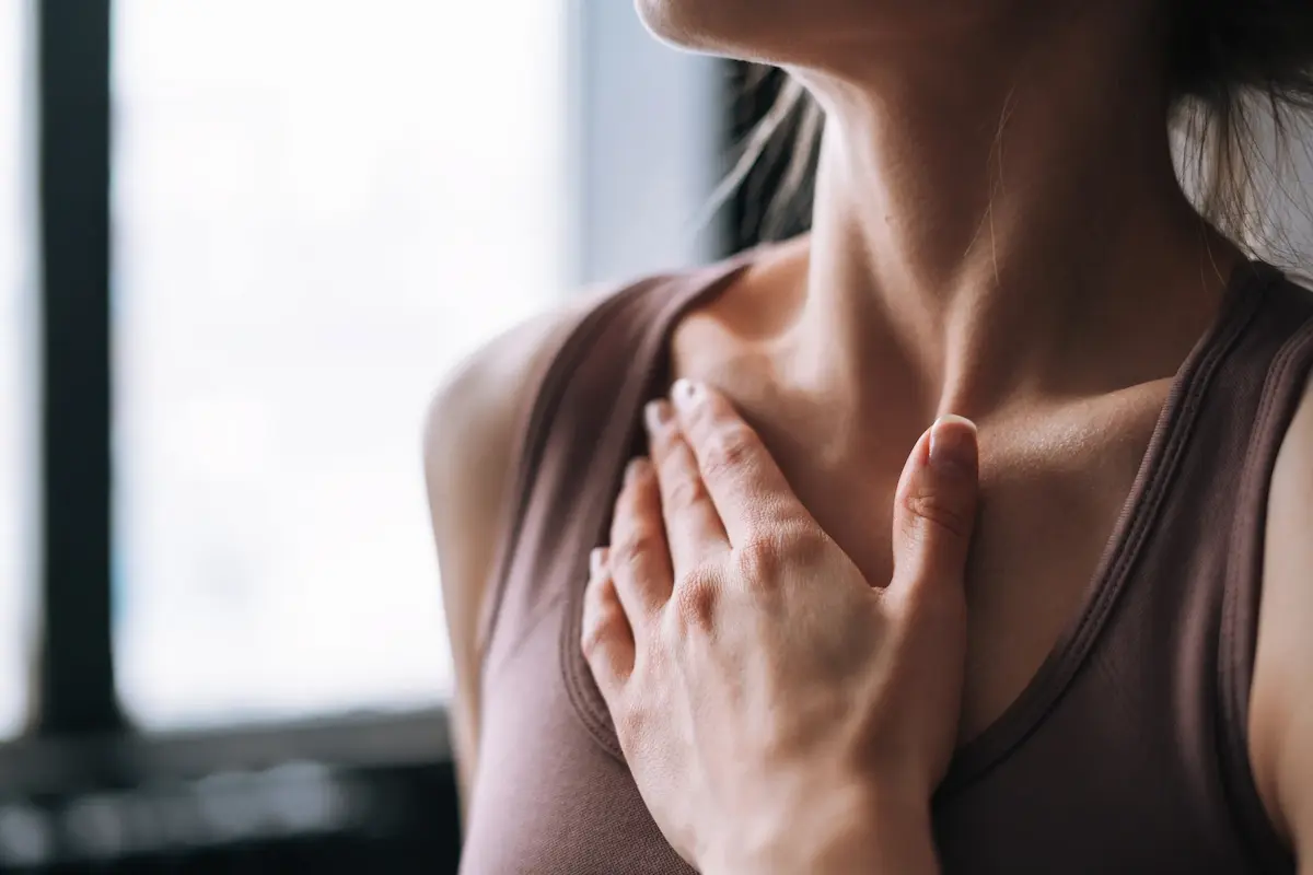 Close-up of a woman's hand resting on her upper chest near the collarbone, wearing a sleeveless top, with a blurred window background.