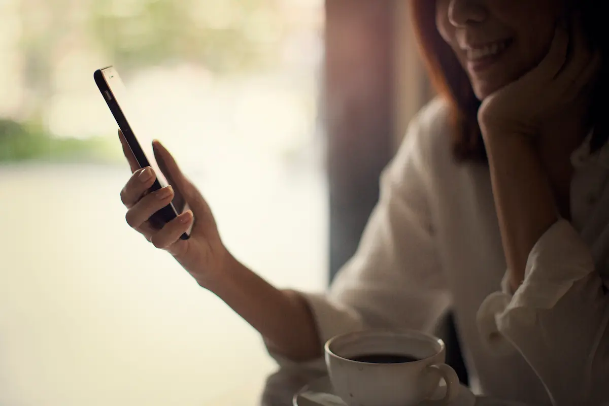 Woman smiling and looking at a smartphone in one hand, sitting at a table with a cup of coffee.